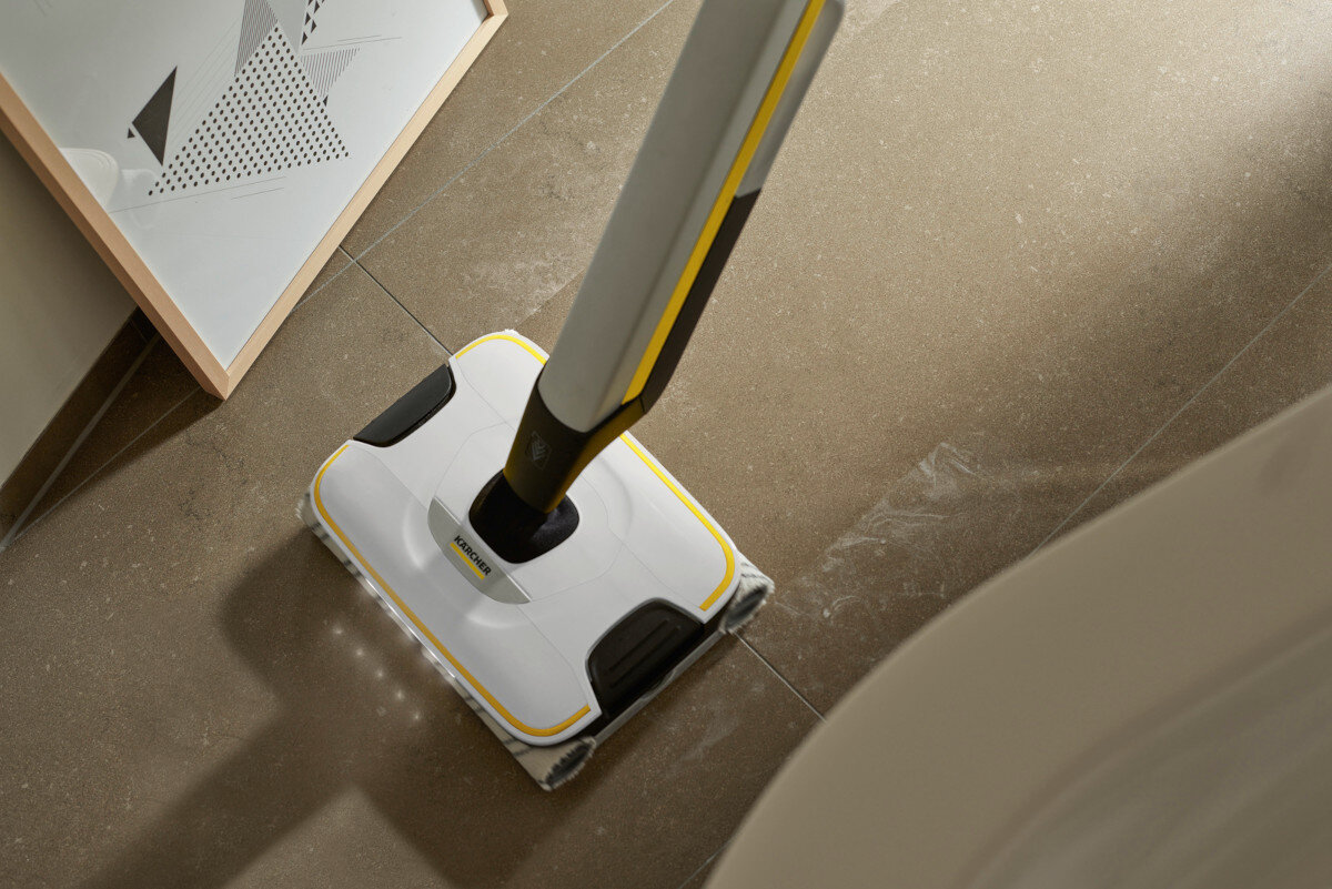 Na obrazie widoczny jest elektryczny mop z logo KARCHER, czyszczący podłogę. Obok znajduje się oparty o ścianę obraz z geometrycznym wzorem.