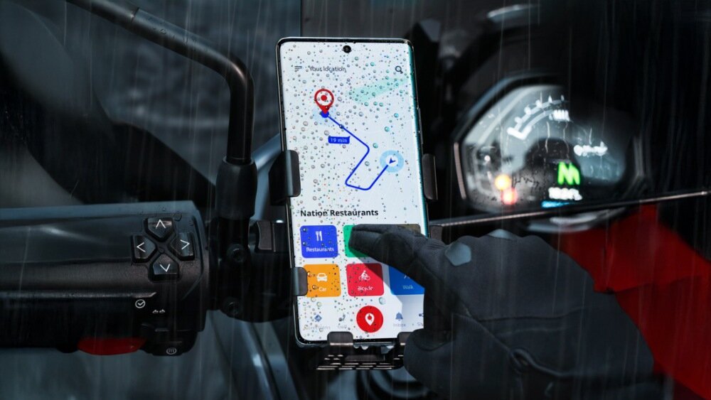 Smartfon zamocowany na kierownicy motocykla wyświetla mapę z trasą i ikonami restauracji. Ekran jest pokryty kroplami deszczu, a osoba w rękawicy dotyka ekranu.