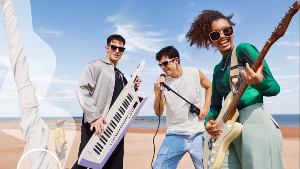 Trzy osoby stoją na plaży, trzymając instrumenty muzyczne: keyboard, mikrofon i gitarę elektryczną. W tle widać morze i niebo z kilkoma chmurami.