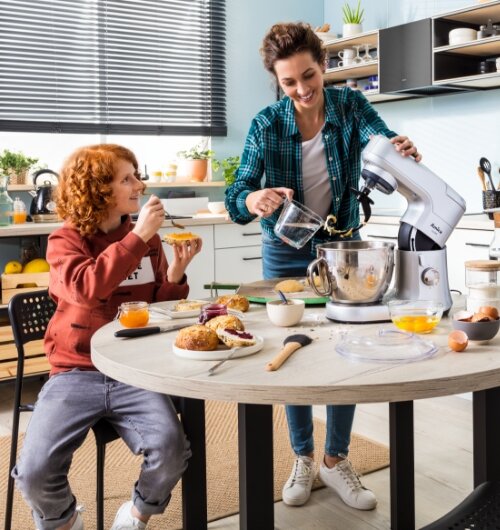 Dwie osoby w kuchni, jedna z nich używa robota kuchennego planetarnego Amica. Na stole znajdują się różne składniki i przybory kuchenne, w tym jajka i pieczywo.