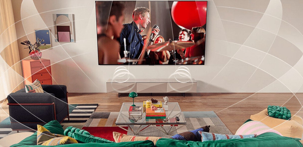 W salonie na ścianie wisi duży telewizor, na którym wyświetlany jest koncert. Przed telewizorem znajduje się kolorowa kanapa i stolik z książkami oraz dekoracjami.