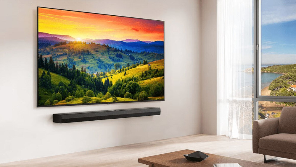 Na ścianie wisi duży telewizor wyświetlający krajobraz z górami i lasem o zachodzie słońca. Pod telewizorem znajduje się listwa dźwiękowa, a obok widoczne jest okno z widokiem na morze i plażę.