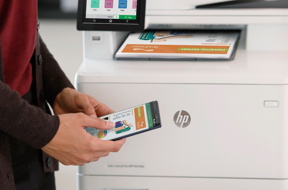 Osoba trzyma smartfon przed urządzeniem wielofunkcyjnym z widocznym logo HP. Na ekranie urządzenia wyświetlane są ikony związane z kopiowaniem i drukowaniem, a w podajniku znajduje się kolorowy dokument.