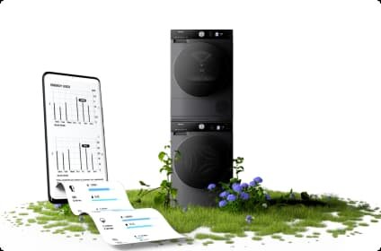Na obrazie widoczna jest czarna suszarka ustawiona pionowo na zielonej trawie z kwiatami, obok której znajduje się smartfon z wykresami i danymi. Suszarka ma nowoczesny design, a na ekranie telefonu widoczne są szczegóły dotyczące zużycia energii.