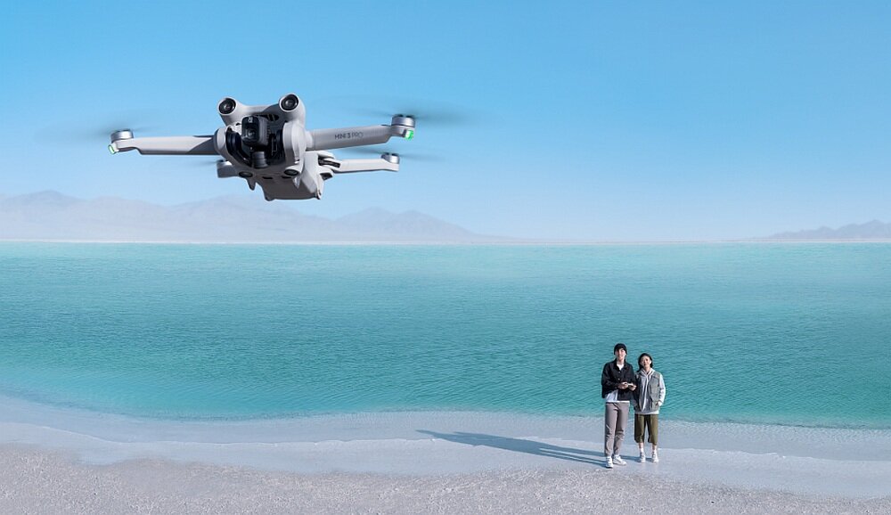 Dron unosi się nad brzegiem morza, a w tle widać dwie osoby stojące na plaży. Niebo jest bezchmurne, a woda ma turkusowy kolor.