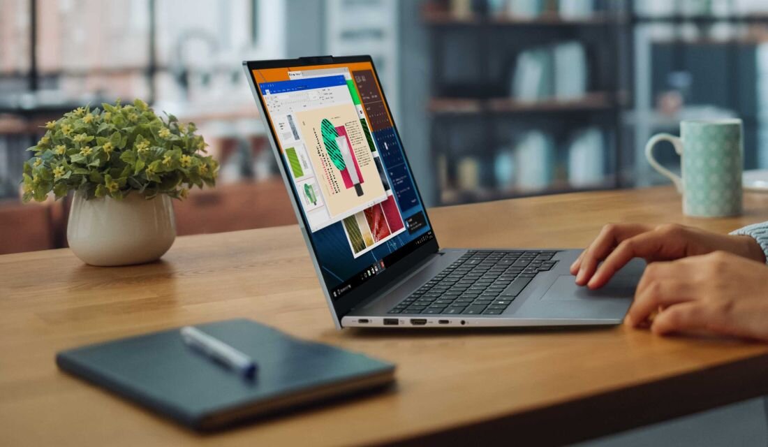 Osoba korzysta z laptopa na drewnianym stole, obok którego znajduje się notes z długopisem i roślina doniczkowa. Na ekranie laptopa widoczne są kolorowe wykresy i dokumenty.