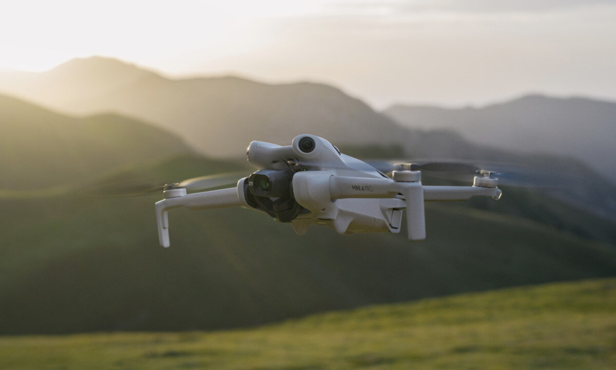 Dron w kolorze jasnym unosi się nad zielonymi wzgórzami przy zachodzącym słońcu. Na dronie widoczny jest napis \'MAVIC\'.