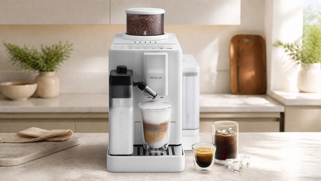 Na blacie kuchennym stoi biały ekspres do kawy z napisem \'DELONGHI RIVELIA\'. Obok ekspresu znajdują się trzy szklanki z różnymi napojami.