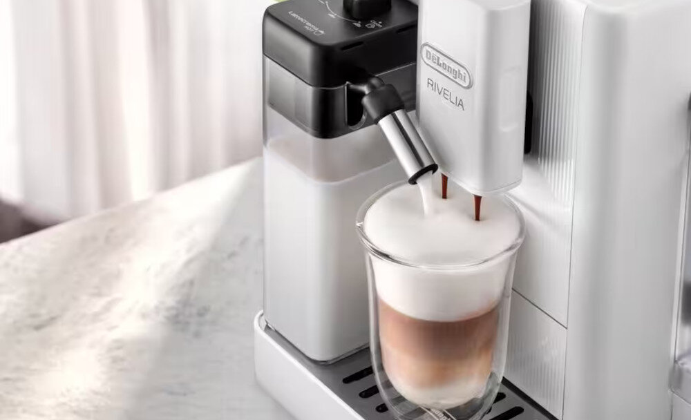Na obrazie widoczny jest ekspres do kawy z logo \'DeLonghi RIVELIA\', przygotowujący kawę z mleczną pianką w szklance. Obok znajduje się pojemnik na mleko z czarną pokrywką.
