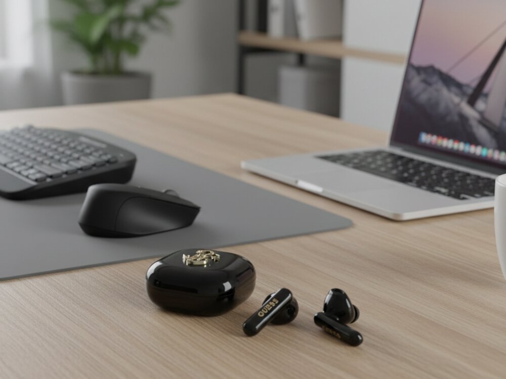 Na drewnianym biurku znajdują się czarne słuchawki douszne z napisem \'GUESS\' oraz czarne etui z metalowym logo. W tle widoczna jest klawiatura, myszka i otwarty laptop.