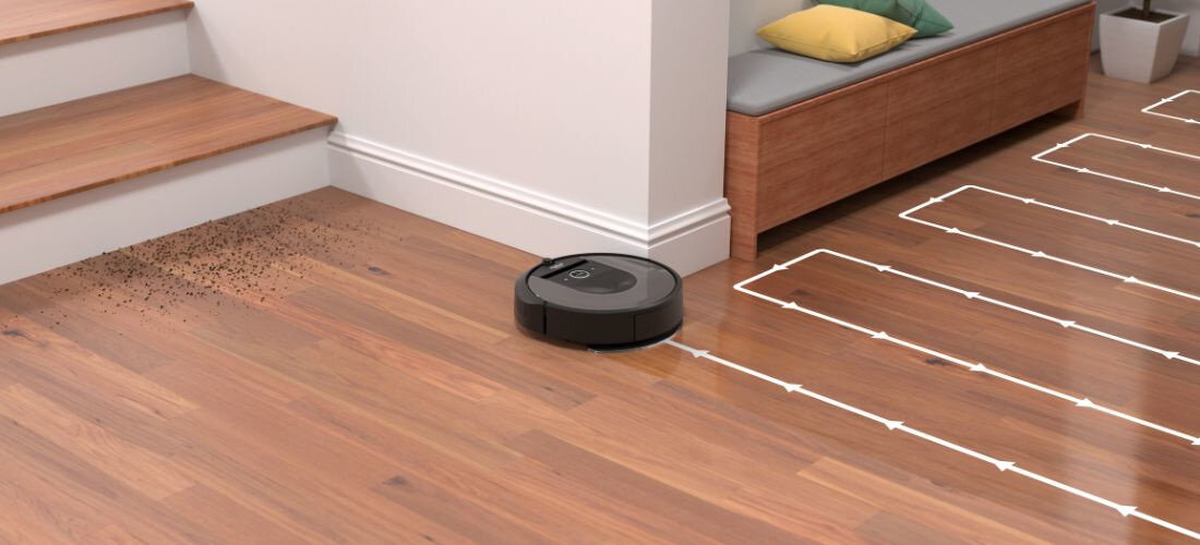 Robot sprzątający porusza się po drewnianej podłodze, zbliżając się do rozsypanych drobinek przy schodach. Na podłodze widoczne są białe linie wskazujące trasę sprzątania.