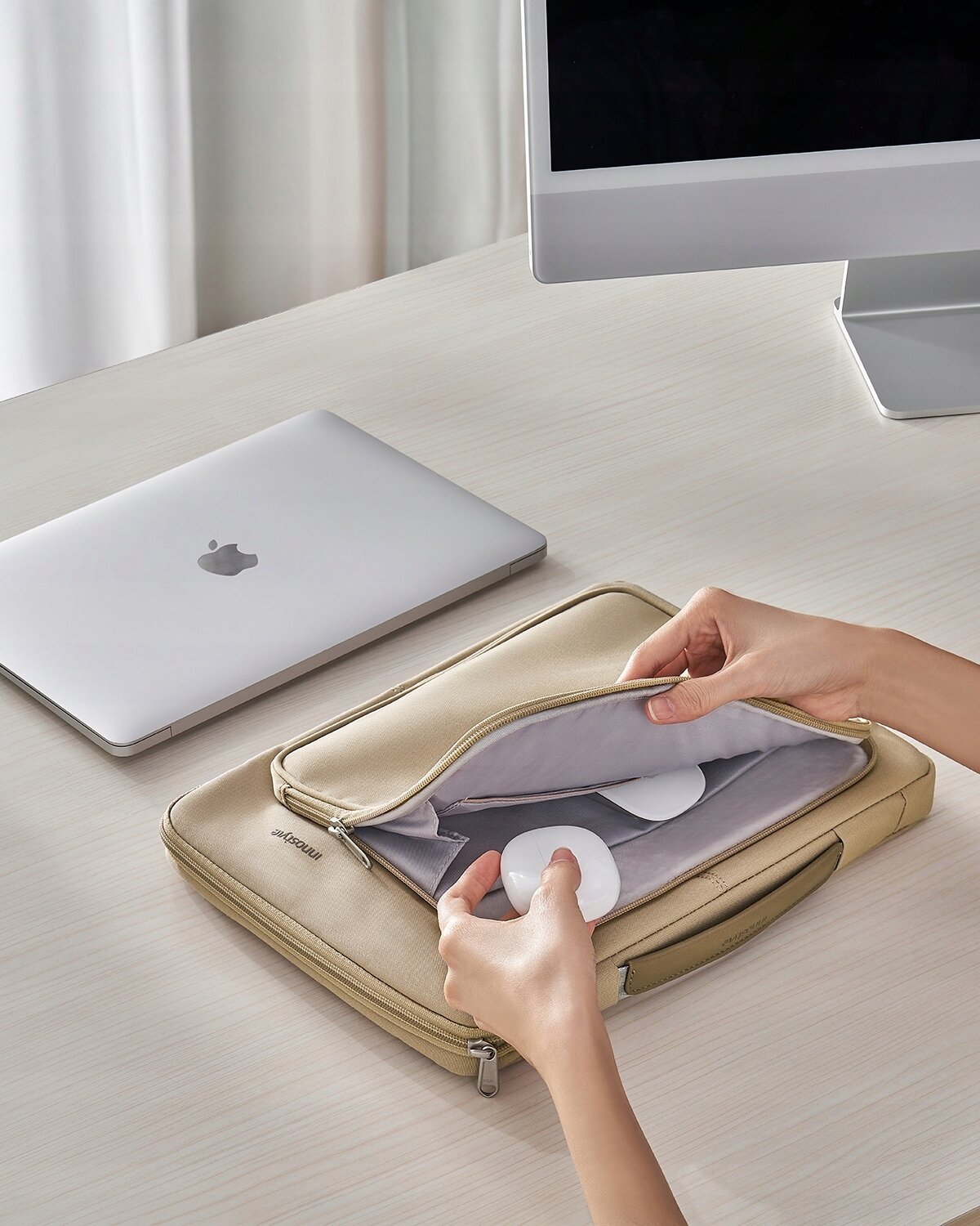 Na zdjęciu widoczna jest beżowa torba na laptopa z otwartą kieszenią, w której znajdują się dwa białe przedmioty. Obok torby leży zamknięty laptop, a w tle znajduje się monitor komputerowy.
