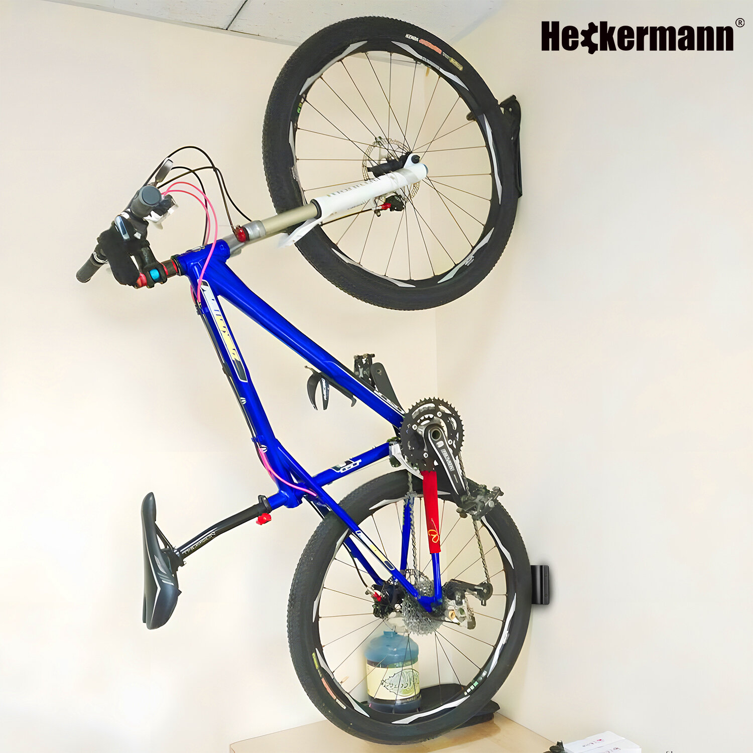 Na ścianie zamontowany jest niebieski rower za pomocą uchwytu ściennego. W prawym górnym rogu widoczne jest logo \'Heckermann\'.