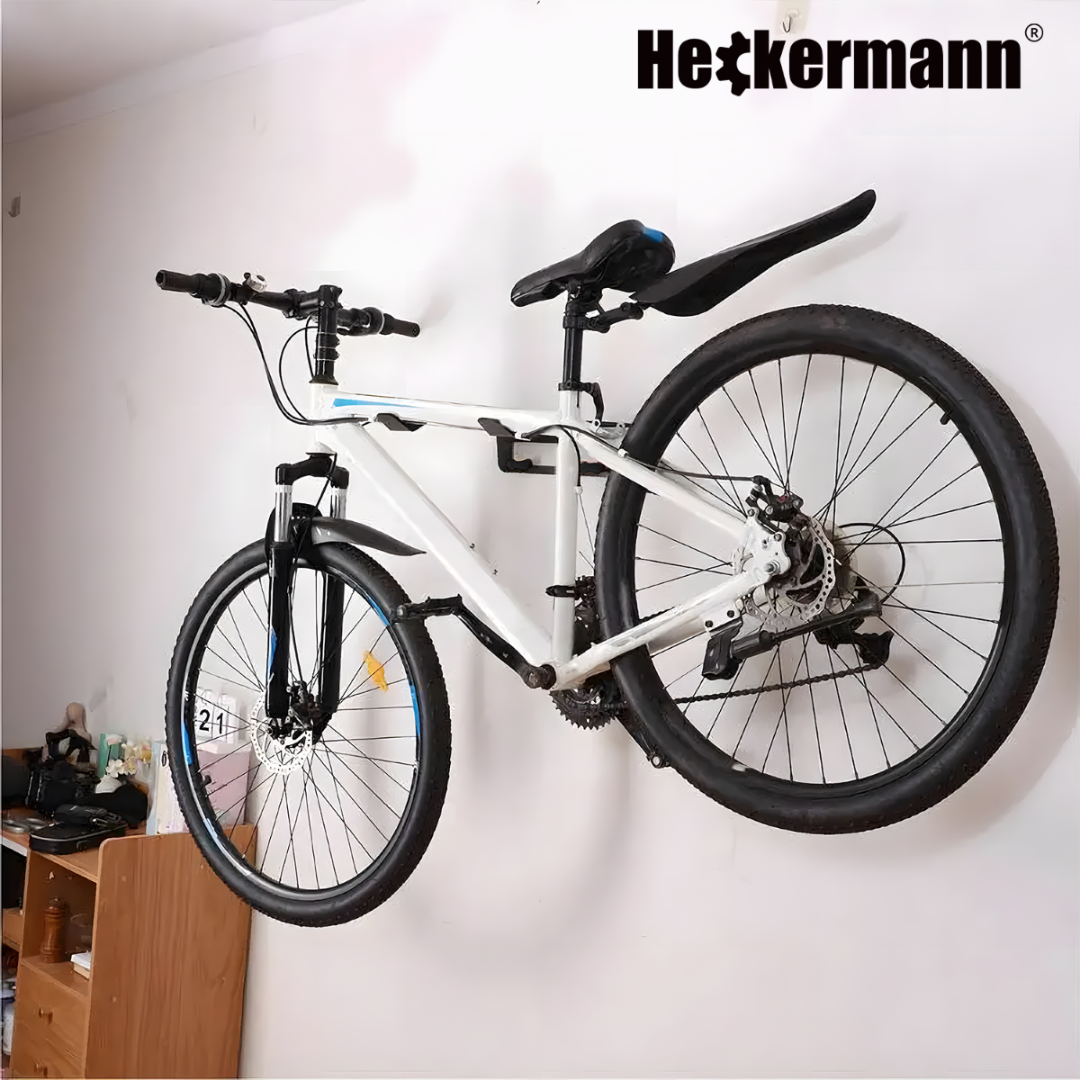 Na ścianie zamontowany jest biały rower przy użyciu uchwytu ściennego. W prawym górnym rogu widoczne jest logo \'Heckermann\'.
