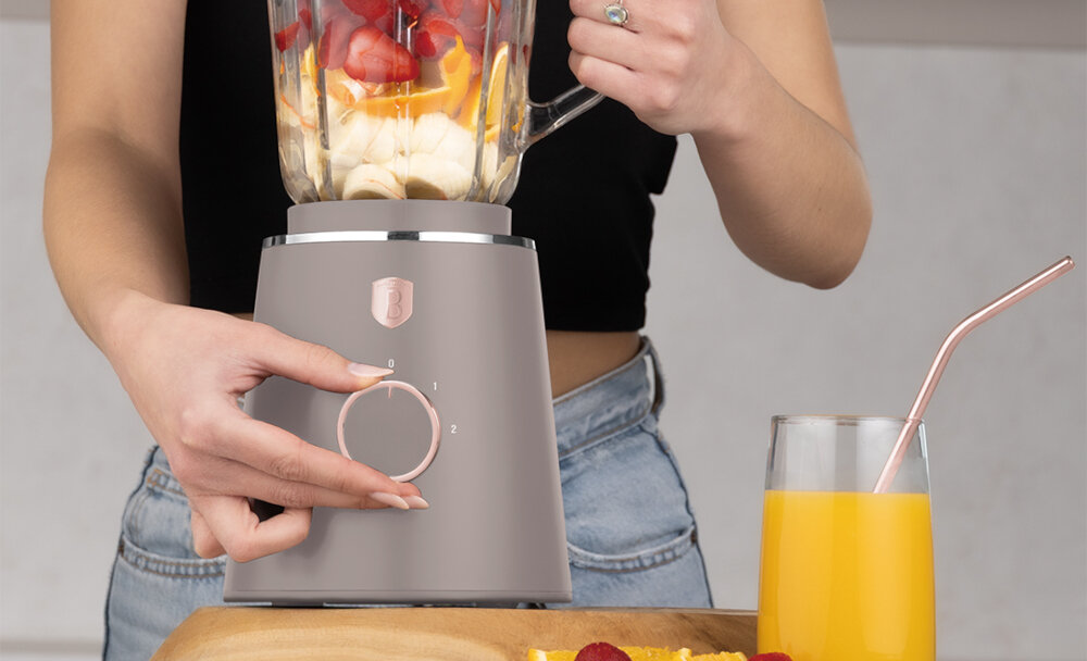 Osoba obsługuje blender kielichowy w kolorze taupe, wypełniony owocami. Obok znajduje się szklanka z sokiem pomarańczowym i metalową słomką.