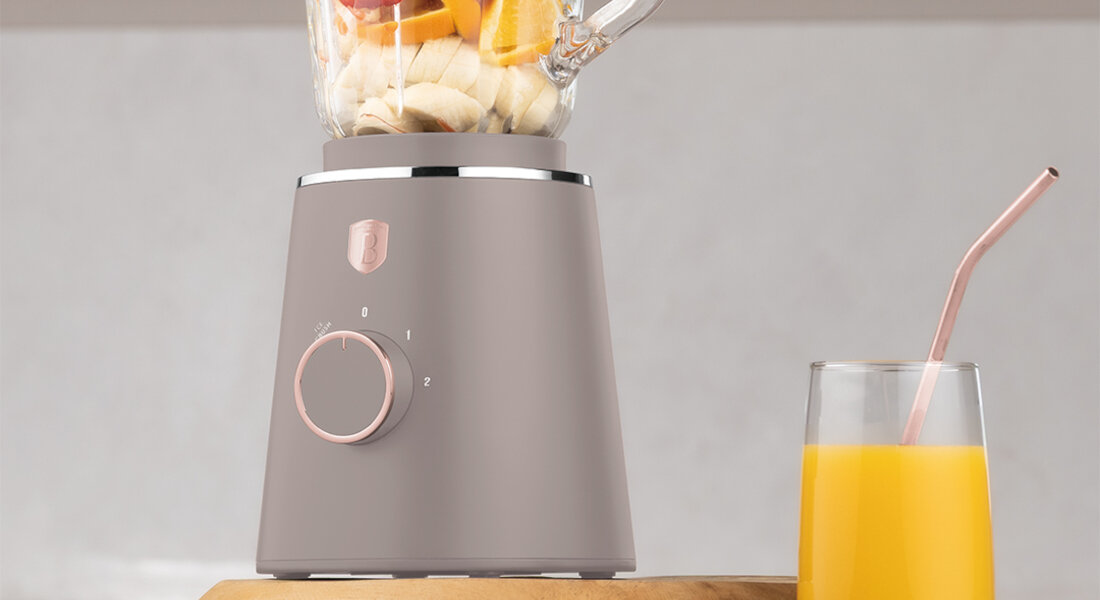 Blender kielichowy w kolorze taupe z pokrętłem i logo producenta, obok szklanka z sokiem pomarańczowym i różową słomką.