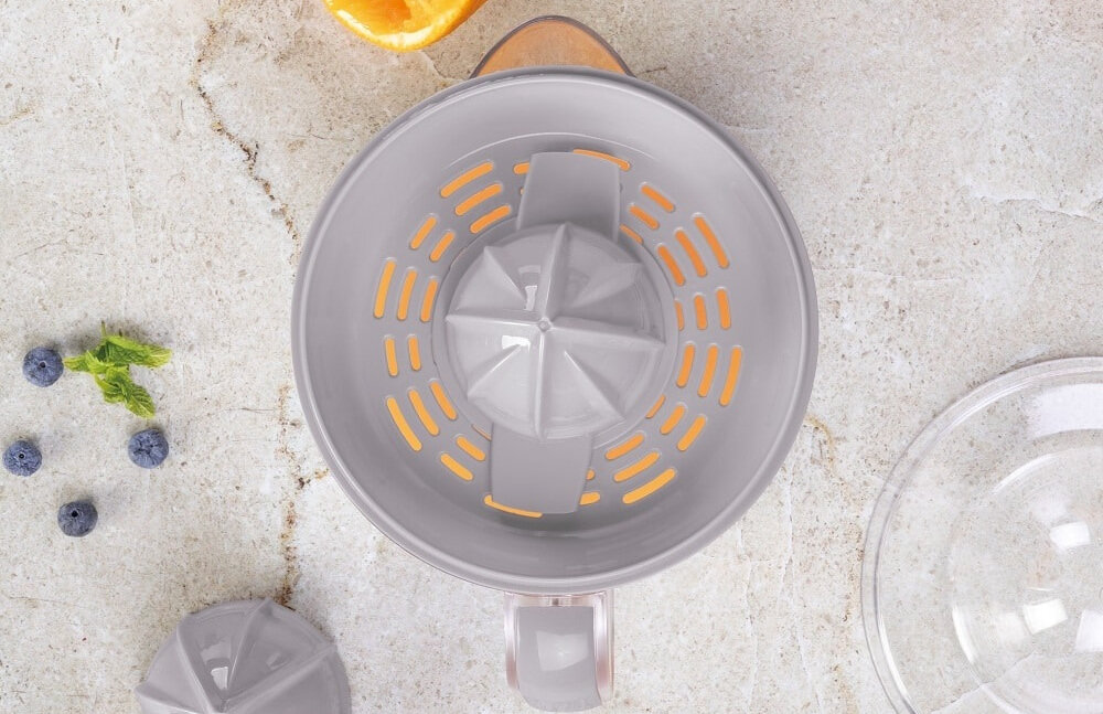 Na obrazie widoczna jest szara wyciskarka do cytrusów z pomarańczowymi elementami wewnętrznymi, umieszczona na jasnym blacie. Obok leżą połówka pomarańczy, kilka jagód i gałązka mięty.