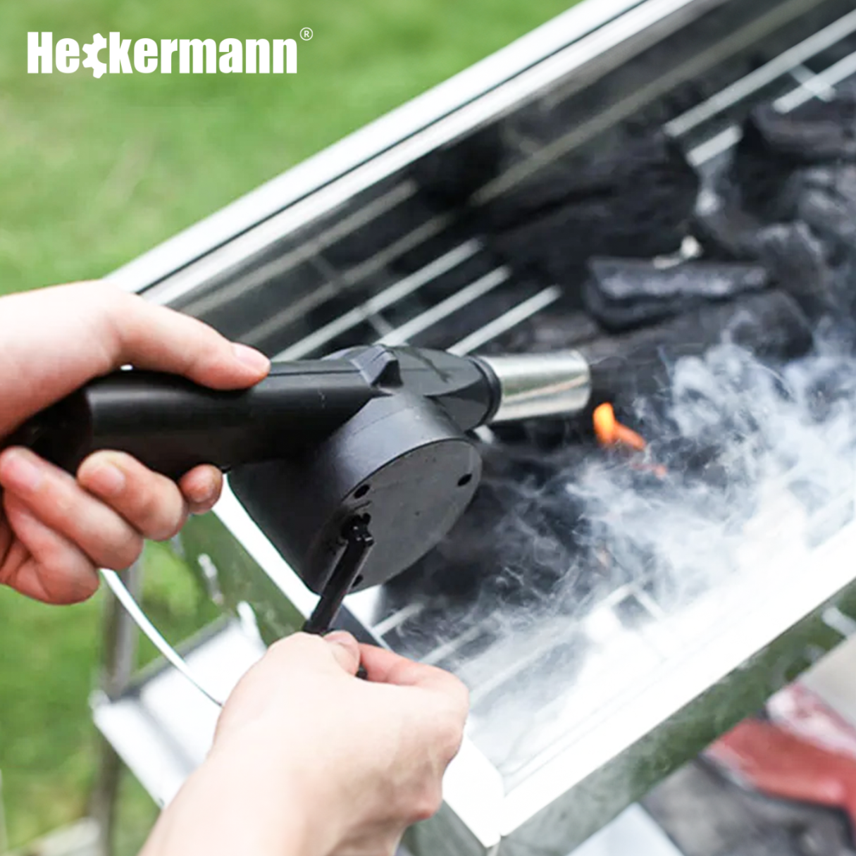 Osoba trzyma ręczny dmuchawę do rozpalania grilla, skierowaną na węgiel drzewny w grillu. W górnym rogu widoczne jest logo \'Heckermann\'.