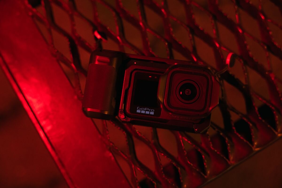 Kamera sportowa z widocznym logo \'GoPro\' leży na metalowej kratce oświetlonej czerwonym światłem. Obiektyw kamery jest skierowany w stronę widza.