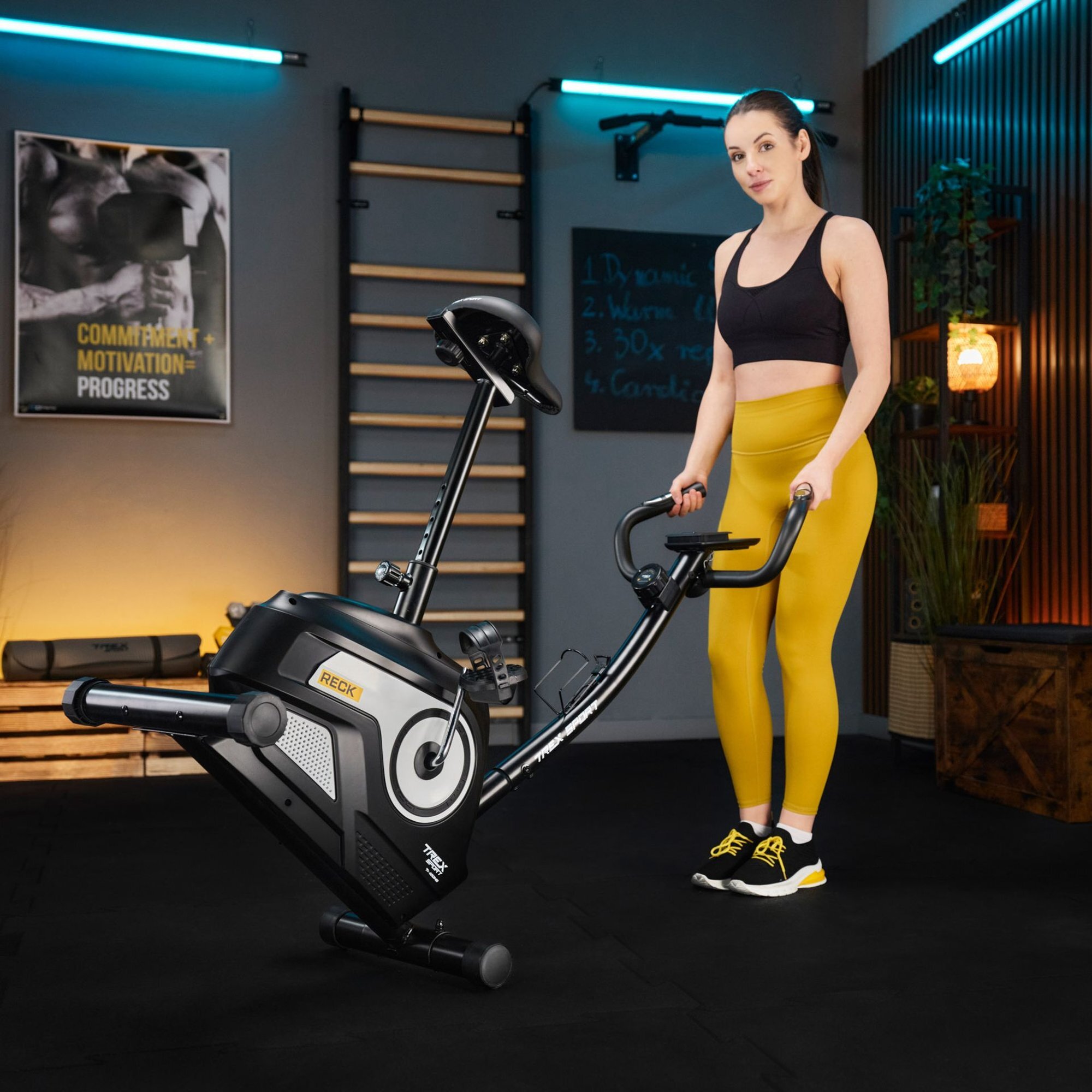Kobieta w sportowym stroju stoi obok roweru stacjonarnego magnetycznego RECK TX-400MB w białym kolorze. W tle widoczne są elementy wyposażenia siłowni oraz plakat z napisem \'COMMITMENT + MOTIVATION = PROGRESS\'.