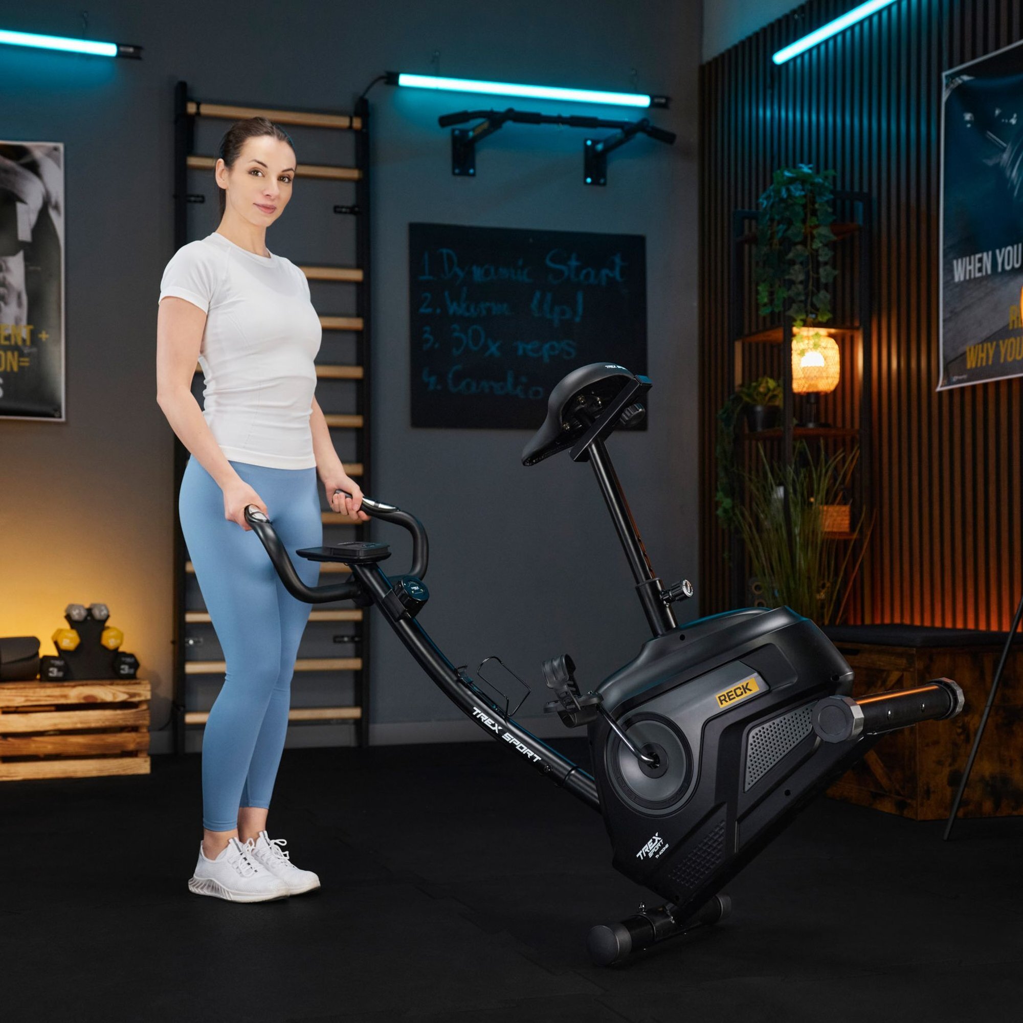 Kobieta stoi obok roweru stacjonarnego magnetycznego RECK TX-400MB w szarym kolorze. W tle widoczne są elementy wyposażenia siłowni oraz tablica z napisem: \'1. Dynamic Start 2. Warm Up! 3. 30x reps 4. Cooldown\'.