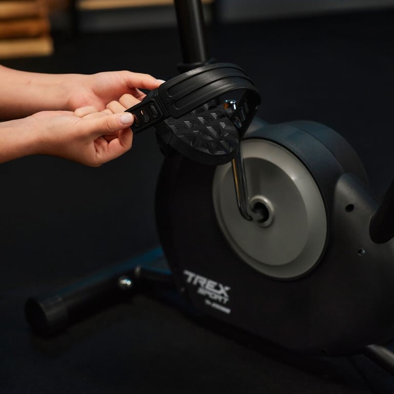 Osoba reguluje pasek pedału roweru stacjonarnego. Na obudowie widoczne jest logo \'TREX Sport\'.