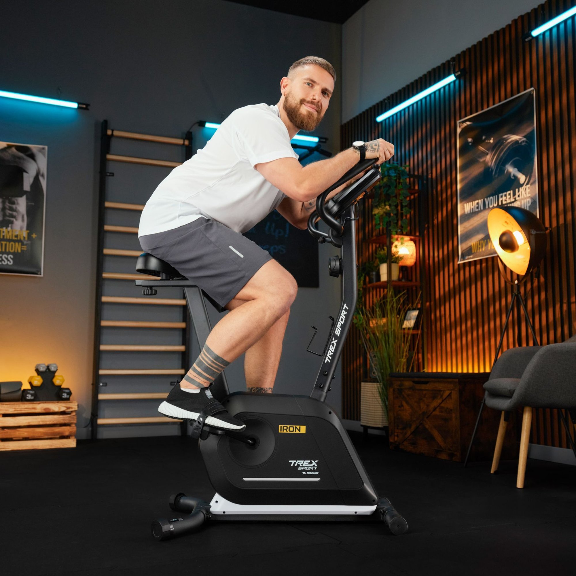 Mężczyzna ćwiczy na czarnym rowerze stacjonarnym magnetycznym IRON TX-500MB w pomieszczeniu z drewnianymi panelami i plakatami na ścianach. Na rowerze widoczne są napisy: \'IRON\' i \'TIREX SPORT\'.