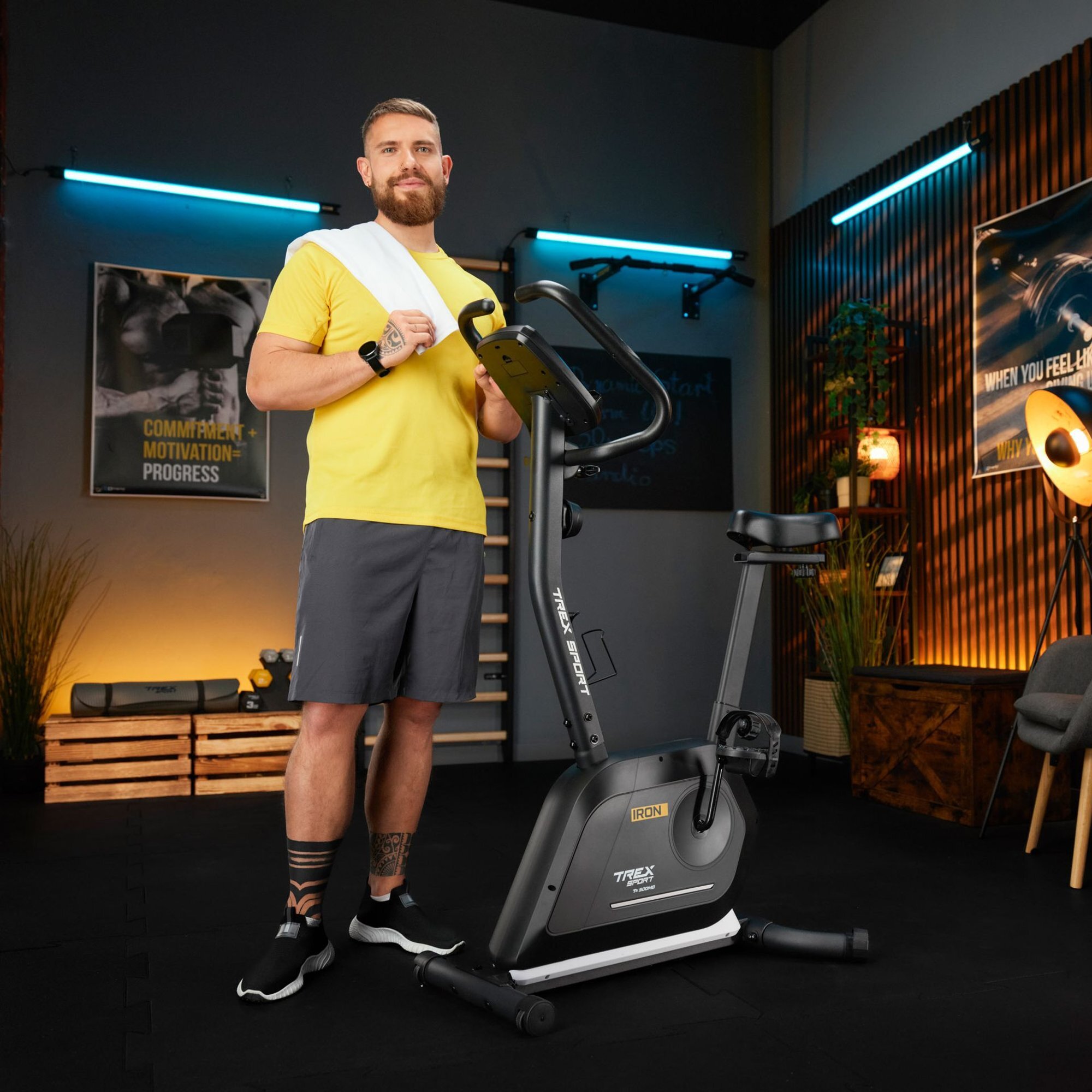 Mężczyzna w żółtej koszulce stoi obok czarnego roweru stacjonarnego z napisem \'IRON\' i \'TREX SPORT\'. W tle widoczne są plakaty i elementy dekoracyjne w pomieszczeniu treningowym.