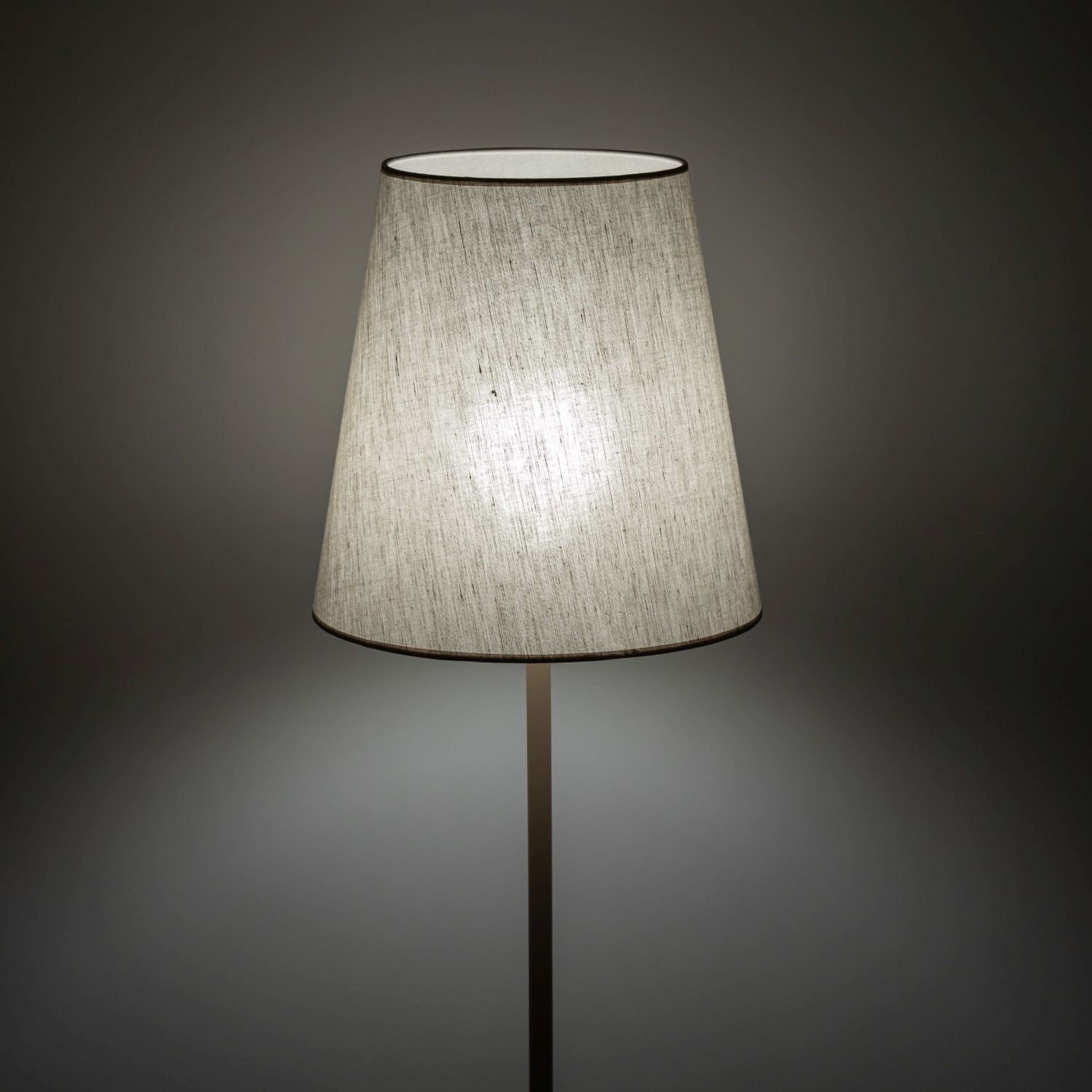 Lampa podłogowa z beżowym, lnianym kloszem na metalowej podstawie. Światło delikatnie przenika przez materiał klosza.