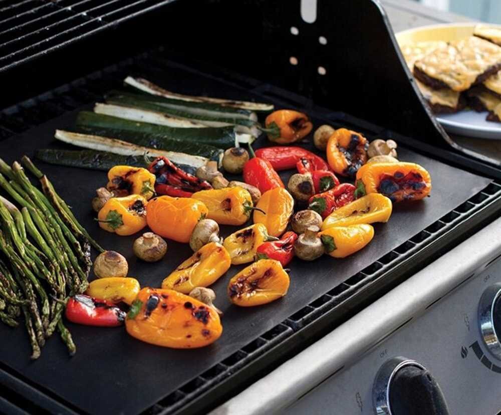 Na macie grillowej znajdują się grillowane warzywa, takie jak papryki, pieczarki, szparagi i cukinia. Obok widać talerz z jedzeniem.