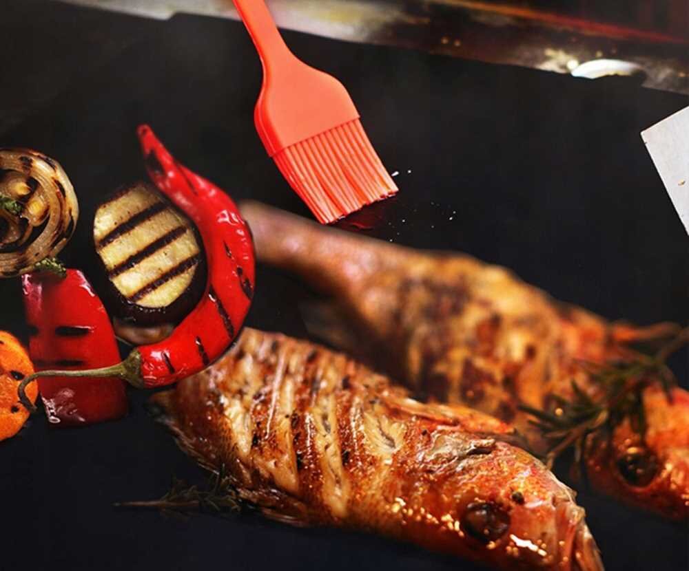 Na obrazie widać grillowane ryby oraz warzywa, takie jak papryka i bakłażan, na czarnej powierzchni. Nad nimi znajduje się czerwona szczotka do smarowania.