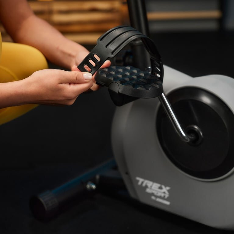 Osoba trzyma pedał roweru stacjonarnego z regulowanym paskiem. Na obudowie roweru widoczne jest logo \'TREX sport\'.
