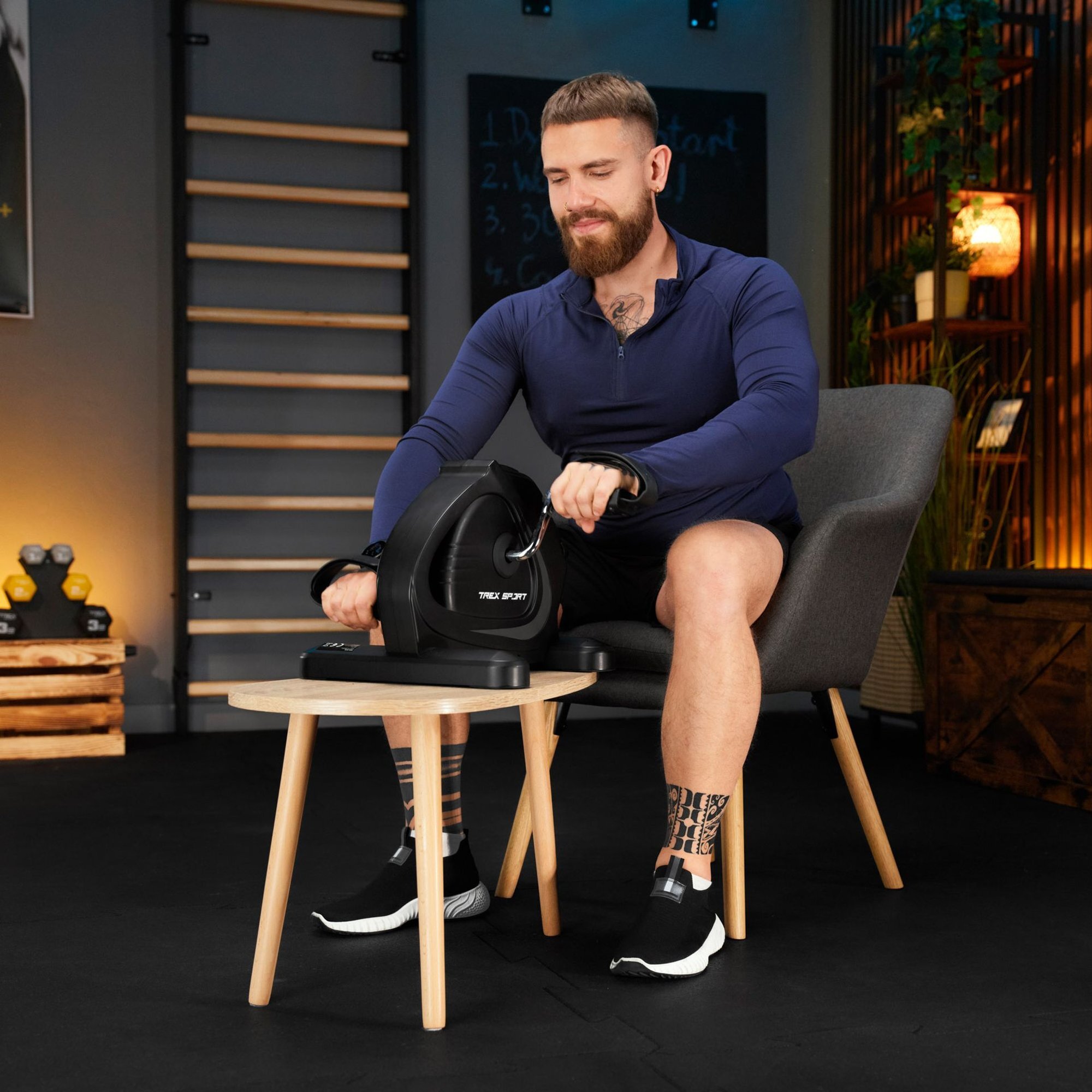 Mężczyzna siedzi na krześle i używa mini roweru treningowego umieszczonego na stoliku. Na urządzeniu widoczne jest logo \'TREX SPORT\'.
