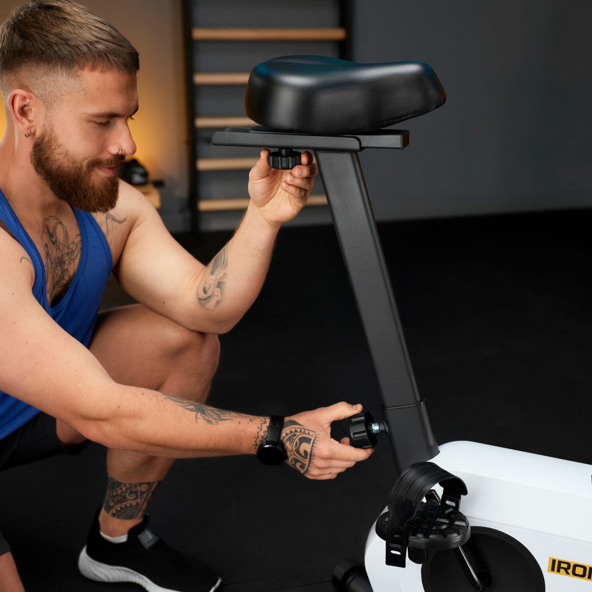 Osoba reguluje wysokość siodełka na białym rowerze stacjonarnym magnetycznym. Na rowerze widoczne jest logo \'IRON\'.