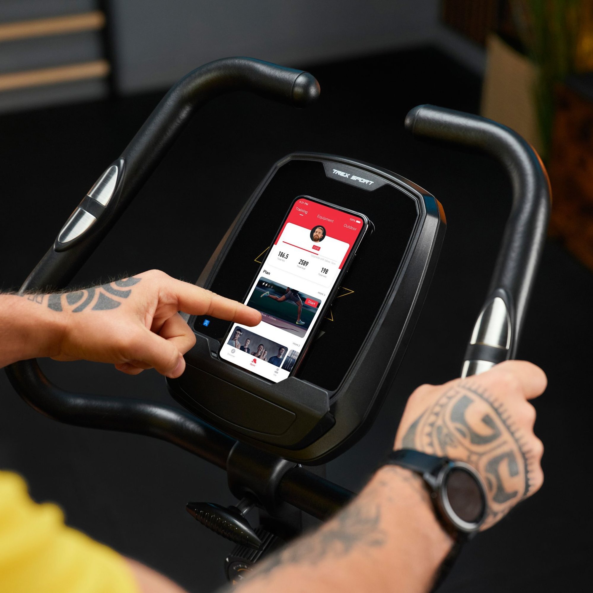Osoba obsługuje panel sterowania roweru stacjonarnego, na którym znajduje się smartfon z aplikacją fitness. Na panelu widoczne jest logo \'TREK SPORT\'.