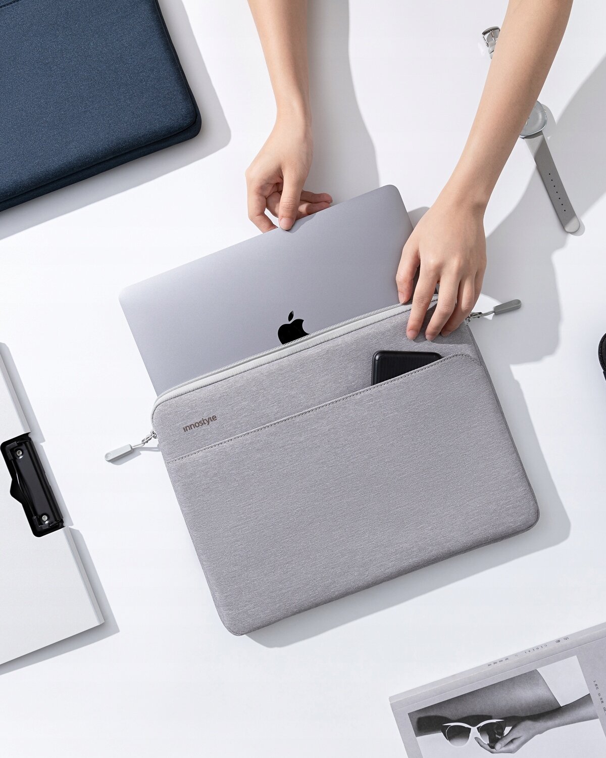 Osoba wkłada laptopa do szarej torby na laptopa z napisem \'innostyle\'. Na torbie znajduje się również kieszeń z telefonem.