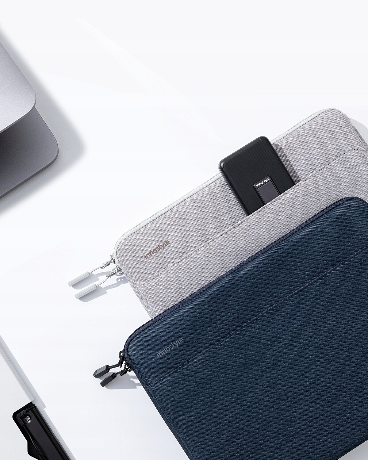 Na obrazie widoczne są dwie torby na laptopa w kolorach szarym i granatowym, z logo \'innostyle\'. Szara torba ma kieszeń, w której znajduje się czarny przedmiot.