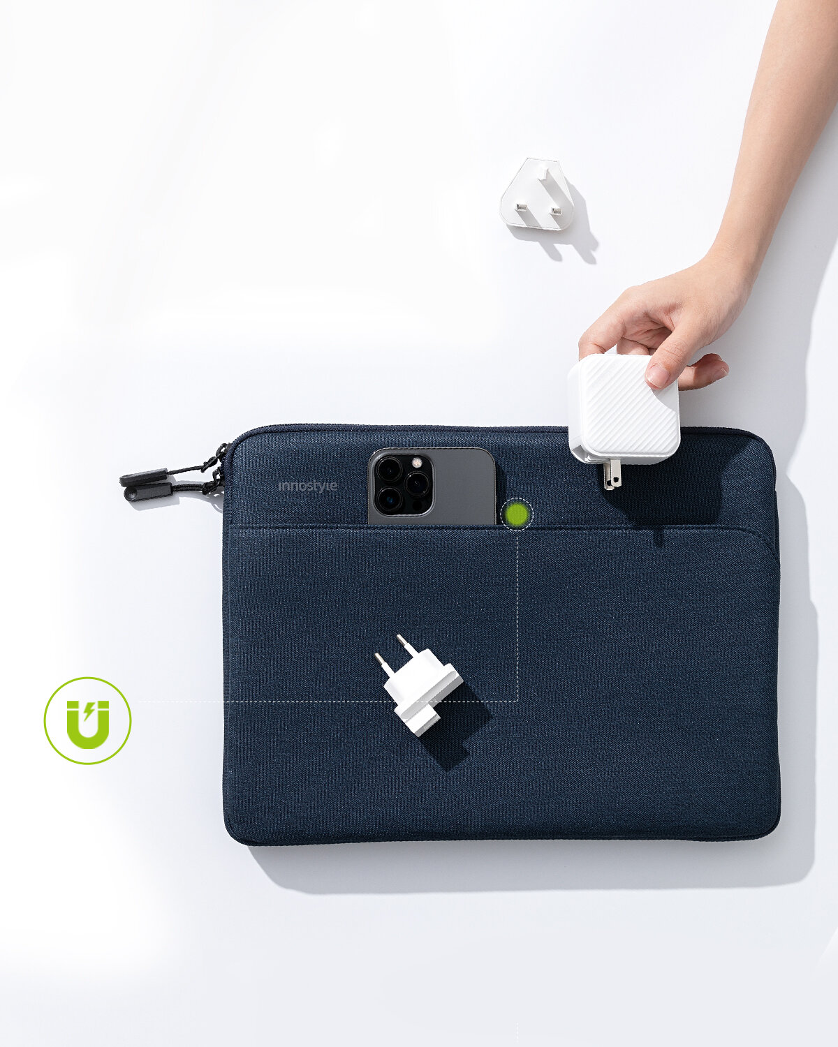 Na obrazie widoczna jest ciemna torba na laptopa z logo \'innostyle\', z której wystaje smartfon i ładowarka. Obok torby znajduje się ręka trzymająca biały adapter oraz dodatkowy adapter leży na powierzchni.