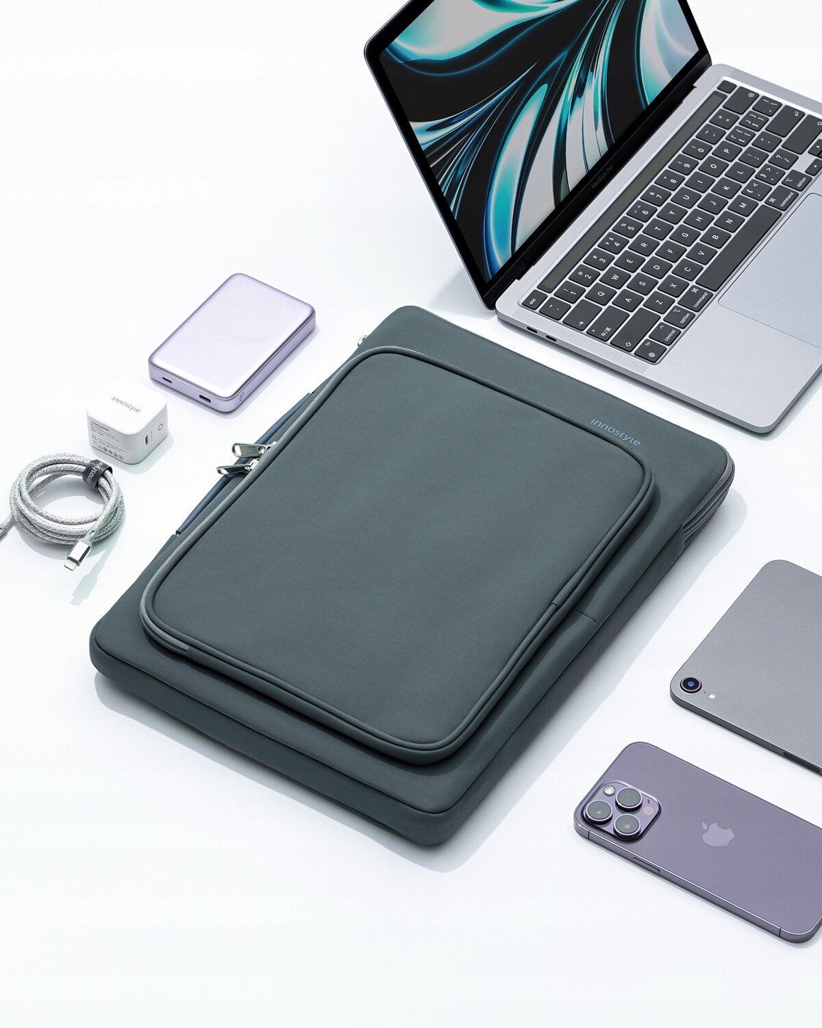 Szara torba na laptopa z napisem \'Innostyle\' leży obok otwartego laptopa, smartfona, kabla, ładowarki i zewnętrznego dysku na białym tle.