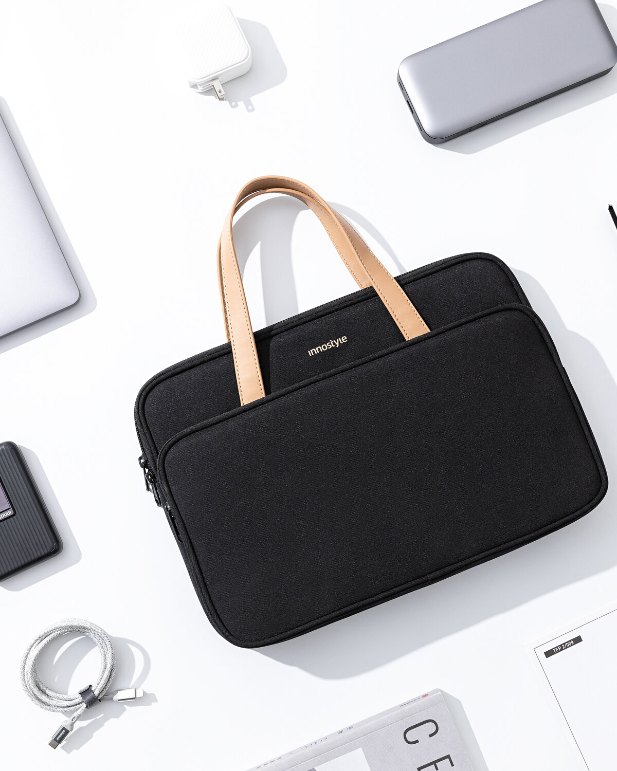 Czarna torba na laptopa z beżowymi uchwytami, z widocznym logo \'innostyle\'. Wokół torby znajdują się różne akcesoria elektroniczne, takie jak ładowarka, kabel i powerbank.