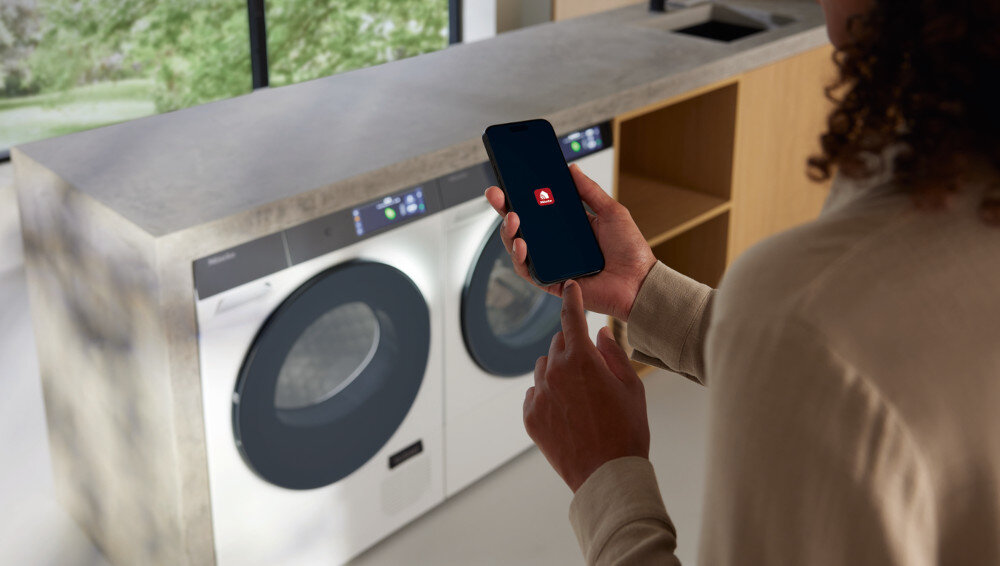 Osoba trzyma smartfon z aplikacją wyświetlającą ikonę pralki. W tle znajdują się dwa urządzenia AGD z okrągłymi drzwiczkami i wyświetlaczami.
