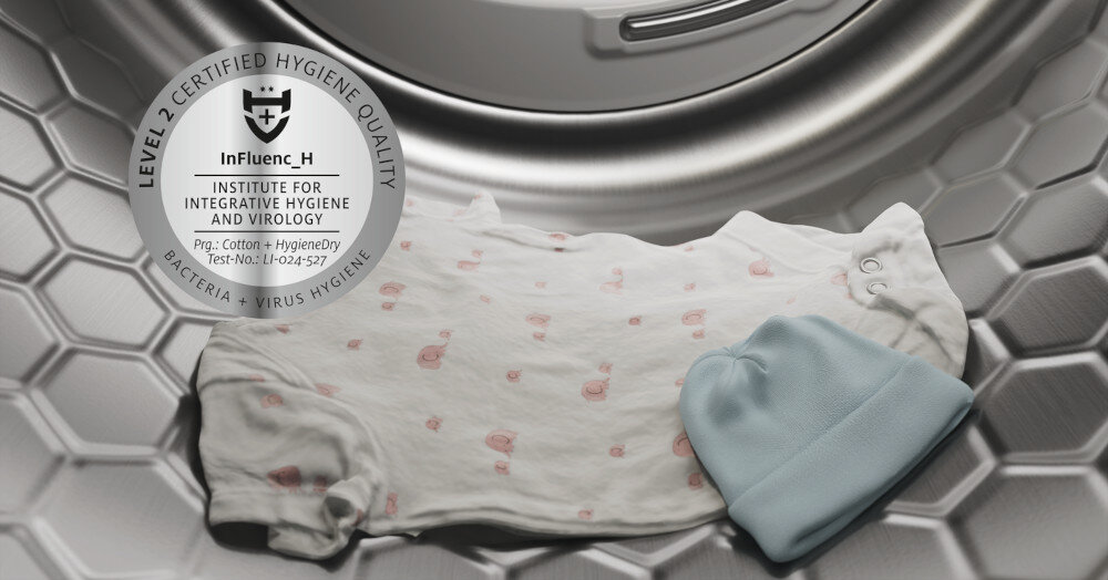 Wnętrze bębna suszarki z białym ubrankiem dziecięcym i niebieską czapką. Widoczny tekst: \'LEVEL 2 CERTIFIED HYGIENE QUALITY, InFluenc_H, INSTITUTE FOR INTEGRATIVE HYGIENE AND VIROLOGY, Prg.: Cotton - HygieneDry, Test-No.: 11-024-527, BACTERIA - VIRUS HYGIENE\'.