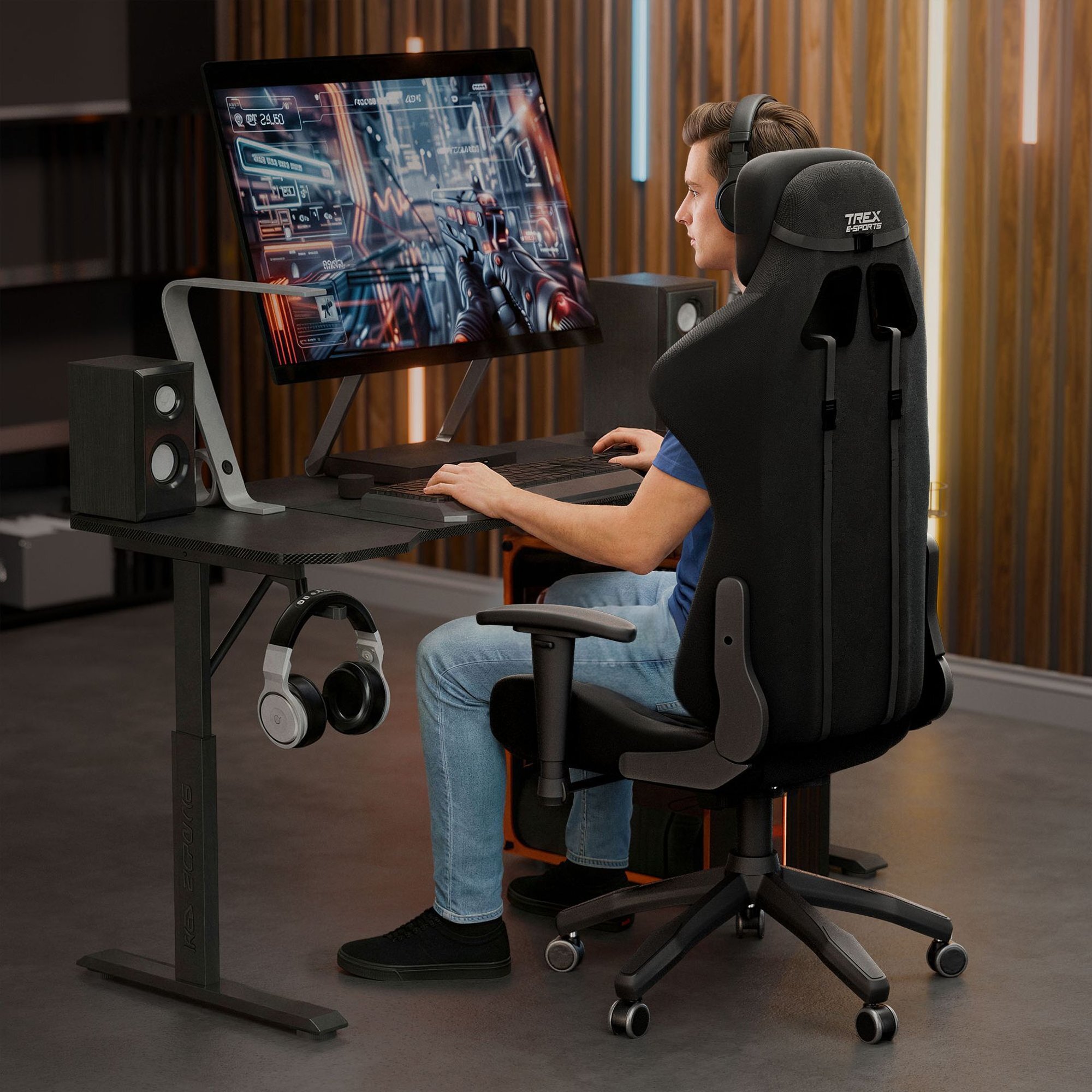 Osoba siedzi na czarnym fotelu gamingowym Trex E-Sports RAVAGE przed biurkiem z dużym monitorem, na którym wyświetlana jest gra. Na biurku znajdują się głośniki, a pod nim wisi para słuchawek.