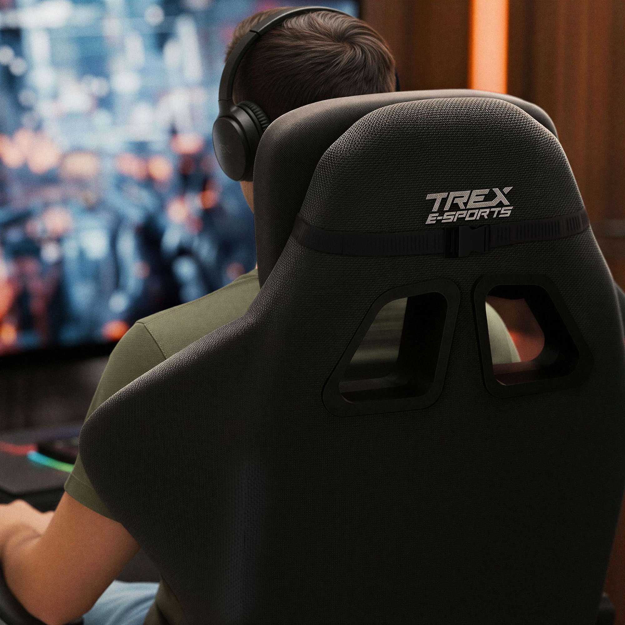 Osoba siedzi na czarnym fotelu gamingowym z widocznym logo \'TREX E-SPORTS\' na oparciu. W tle znajduje się rozmyty ekran z kolorowymi elementami.