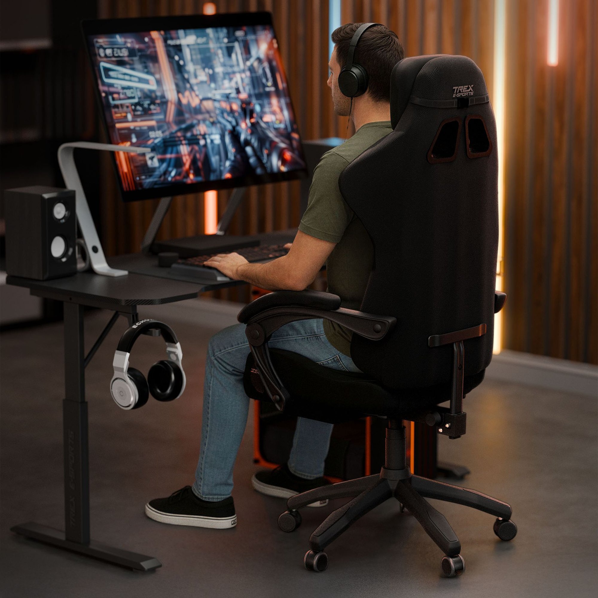 Osoba siedzi na czarnym fotelu gamingowym z napisem \'TREX E-SPORTS\' na oparciu, przed dużym monitorem z wyświetlonymi grafikami. Na biurku znajdują się głośniki, a na uchwycie wisi para słuchawek.