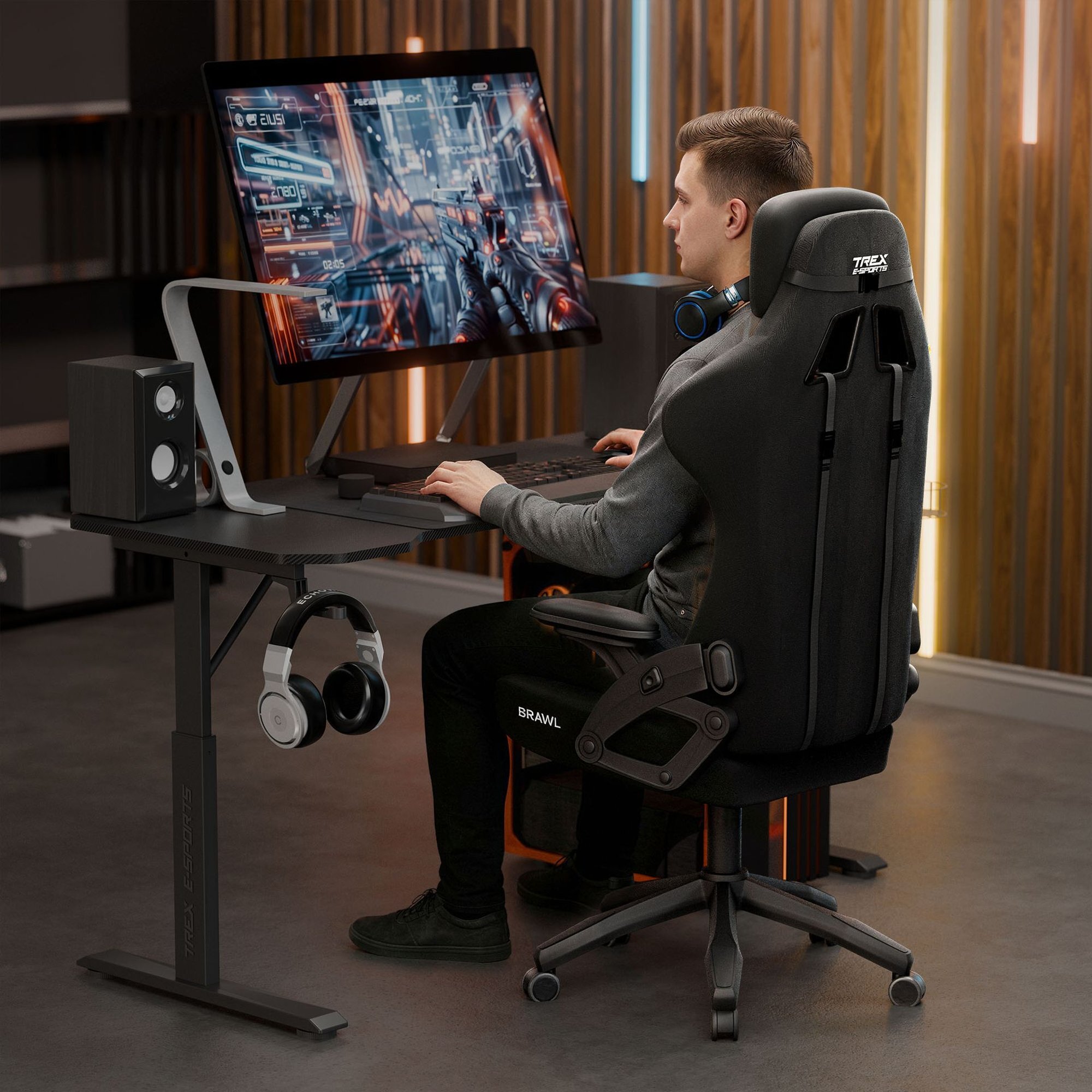 Osoba siedzi na czarnym fotelu gamingowym z napisem \'TREX E-SPORTS\' i \'BRAWL\', przed dużym monitorem z wyświetlonym interfejsem gry. Na biurku znajdują się głośniki i słuchawki, a w tle widoczne są drewniane panele ścienne.