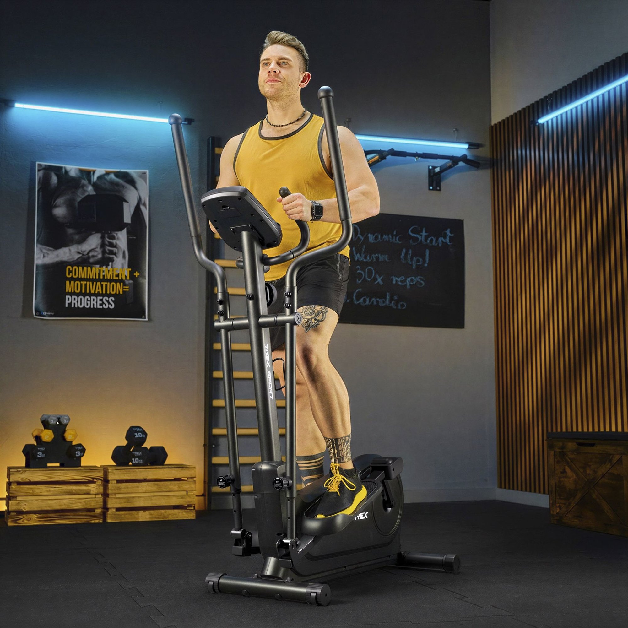 Osoba ćwiczy na czarnym orbitreku magnetycznym w pomieszczeniu z plakatem \'COMMITMENT + MOTIVATION = PROGRESS\'. W tle widoczne są hantle i tablica z napisem \'Dynamic Start Warm Up! 30x reps Cardio\'.