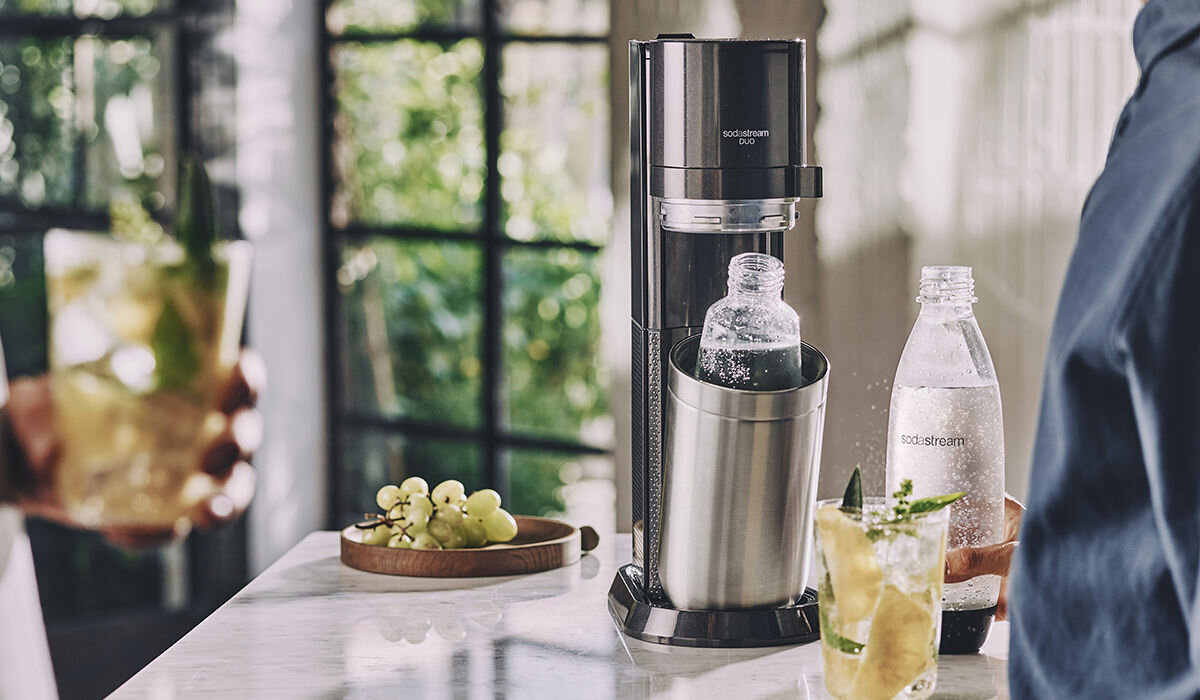 Na blacie kuchennym stoi czarny saturator z napisem \'sodastream duo\', obok którego znajdują się dwie butelki z wodą gazowaną. W tle widać osobę trzymającą szklankę z napojem i talerz z winogronami.