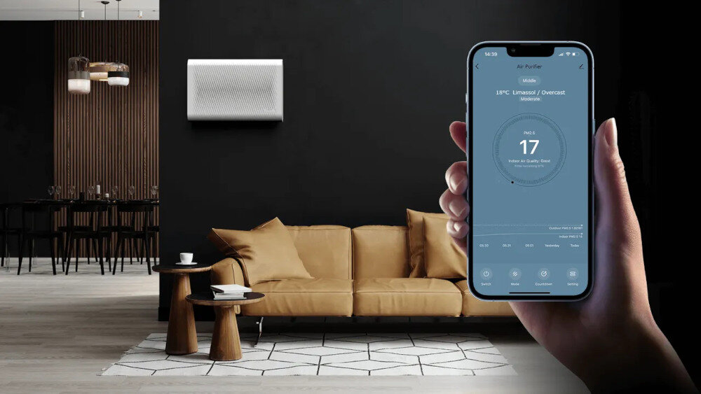 W eleganckim salonie z ciemnymi ścianami i jasnobrązową sofą, na ścianie zamontowany jest oczyszczacz powietrza. Osoba trzyma smartfon z aplikacją pokazującą dane dotyczące jakości powietrza, z wartością \'17\' na ekranie.