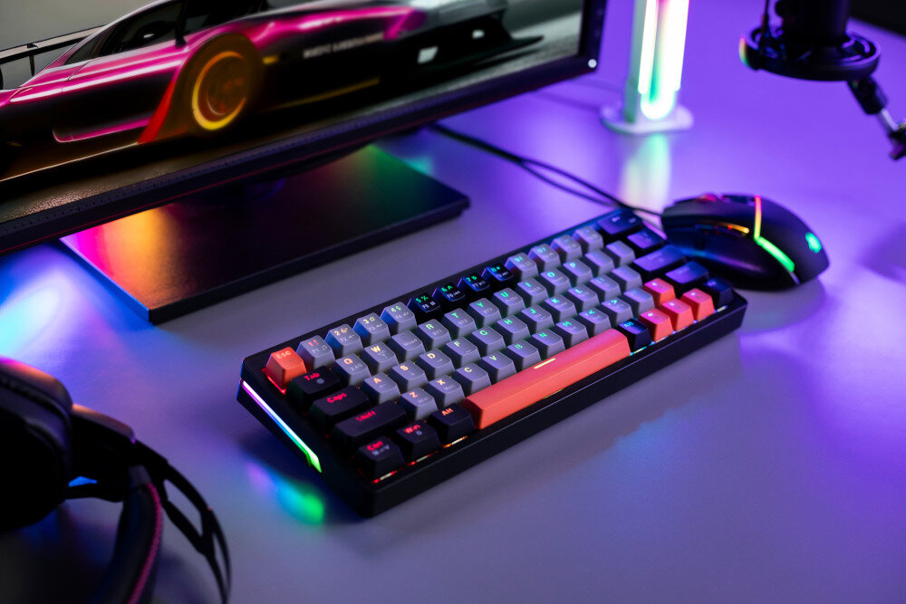 Na biurku znajduje się klawiatura z podświetleniem RGB, obok której leży mysz komputerowa. W tle widać monitor z wyświetlonym obrazem samochodu wyścigowego oraz słuchawki i mikrofon.